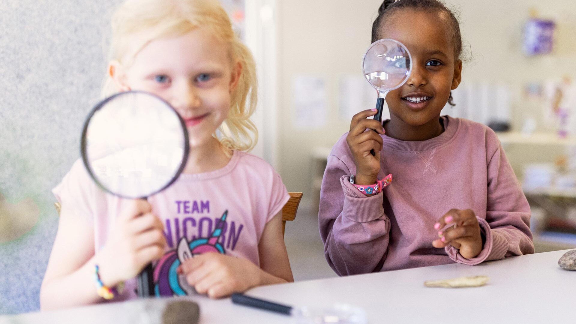 Två barn på förskolan sitter och tittar in i kameran med varsitt förstoringsglas.